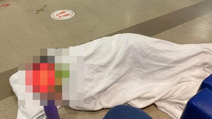 A person covered with a white blanket lays on a hospital floor, their face is blurred