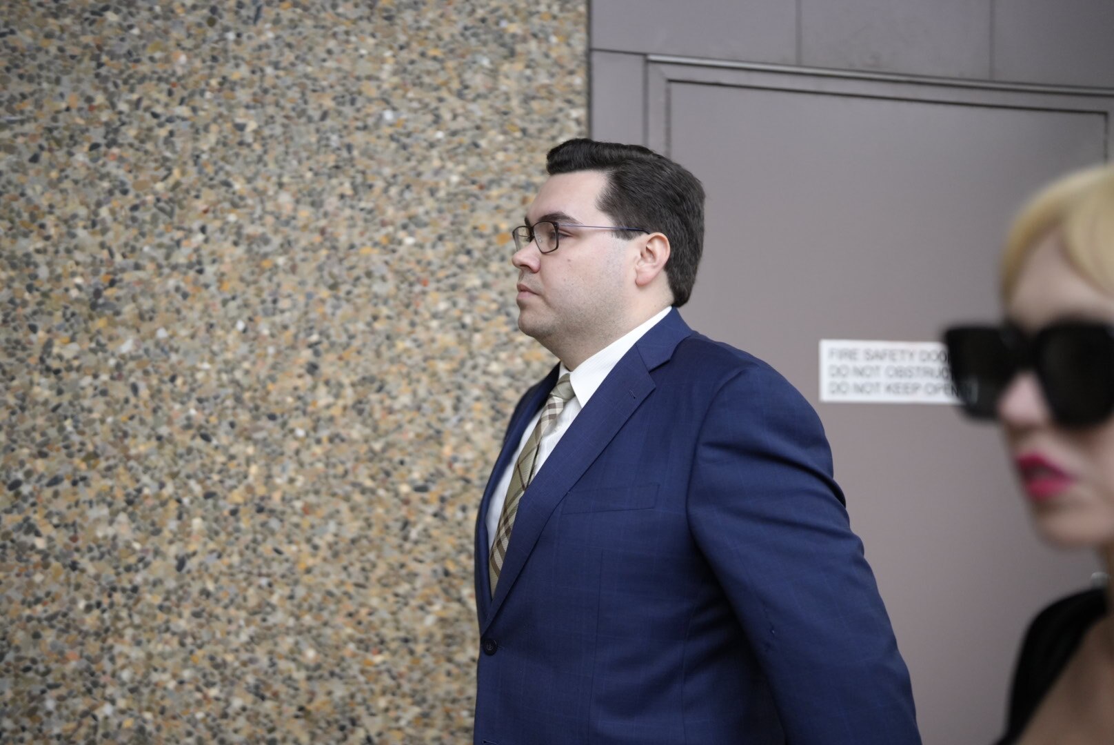 Un hombre con pelo corto y oscuro y gafas, vestido con un traje azul oscuro, camina, visto desde un lado.