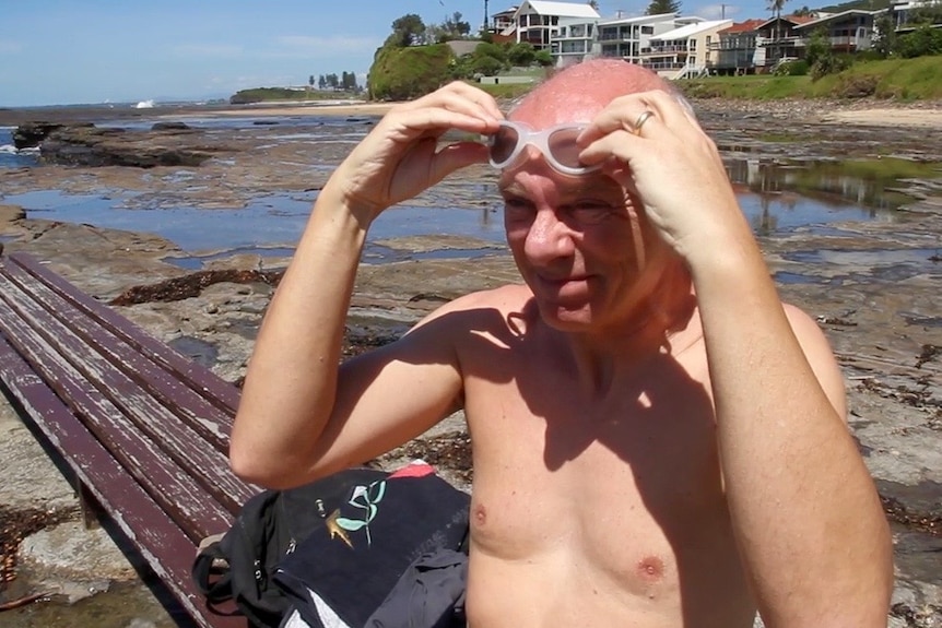 A man sits on a bench by the coastline, pulling his goggles on.