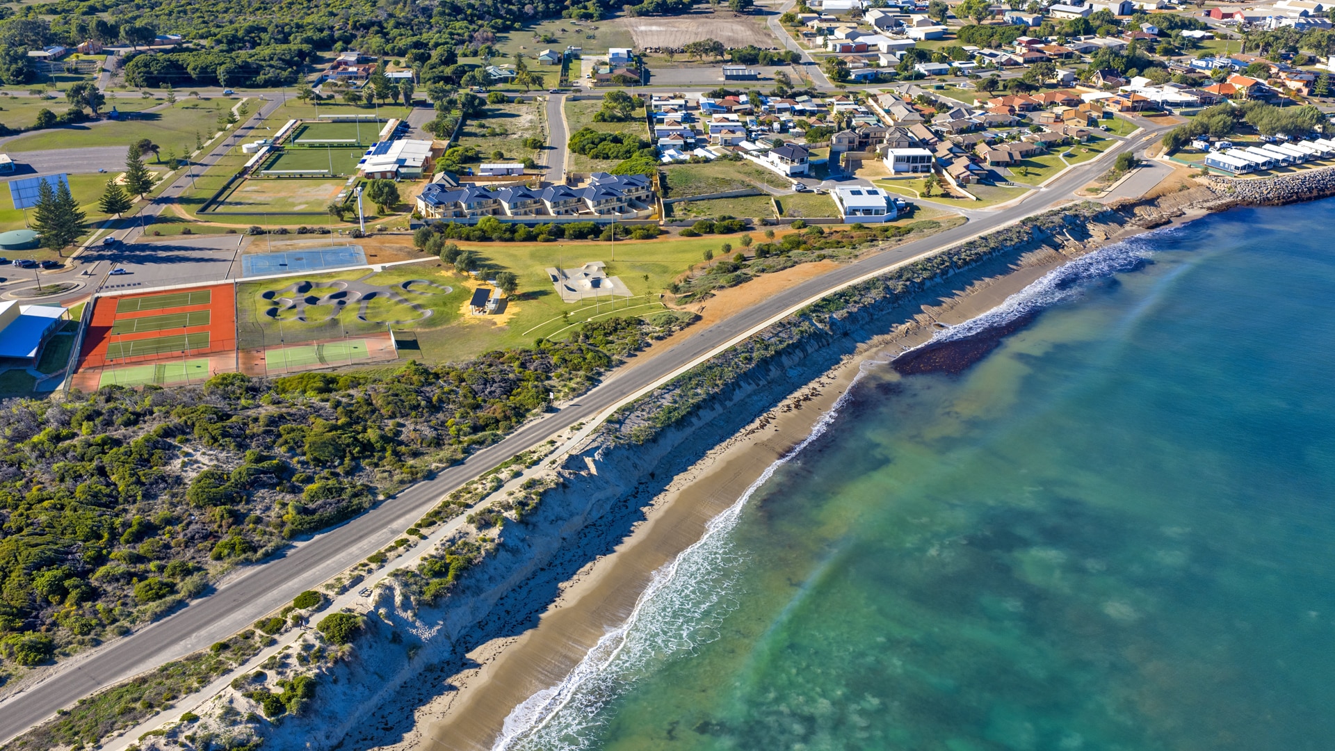 A drone shot shows the ocean and erosion near a road. Behind it sits a recreation centre, playground, bike track and houses.