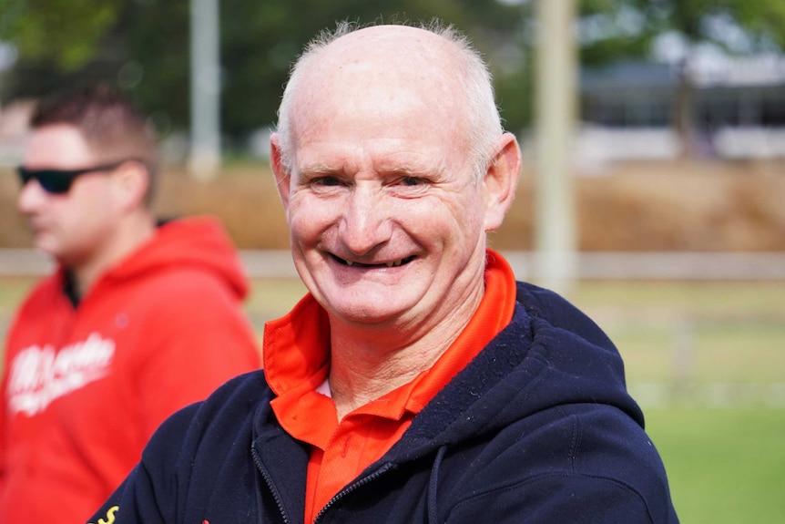 Coach Keith Seiler in a red polo shirt and navy hooded jumper smiles for a photo on a soccer pitch.