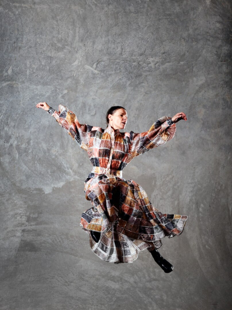 A dancer in a flowing patchwork dress leaps into the air with her arms outstretched,