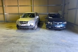 A silver four-wheel-drive and a blue sedan are pictured inside a garage. Each has minor damage.
