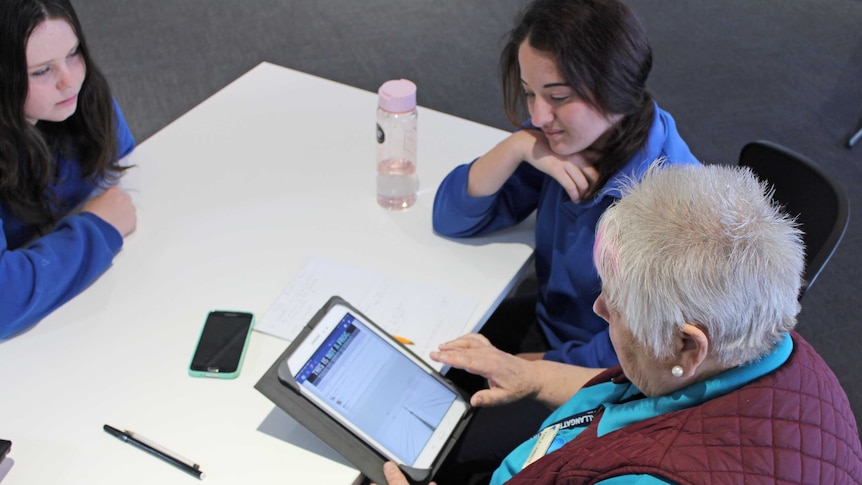 Two female high school students help a 60-year-old woman use Facebook on her ipad