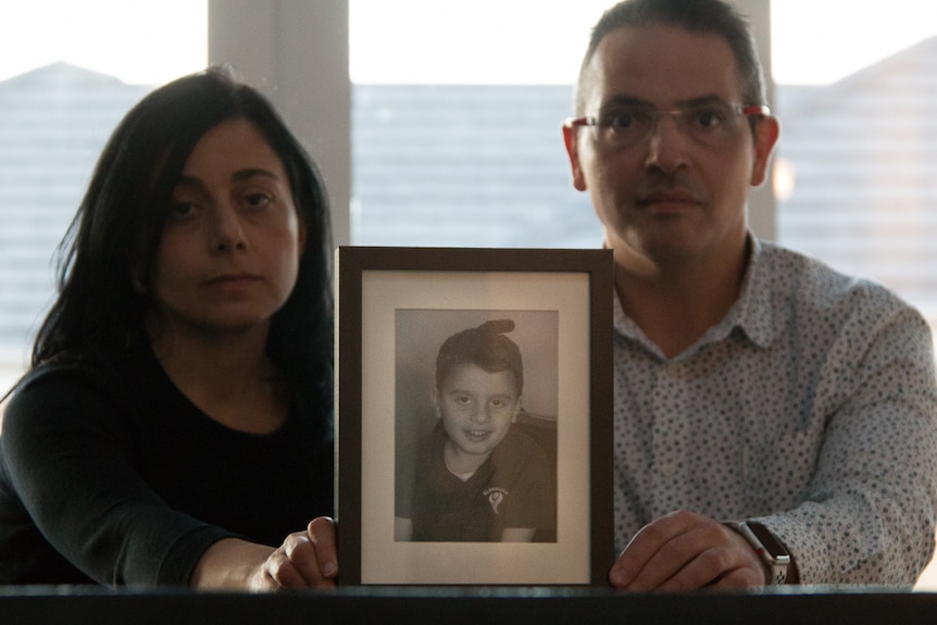 A man and a woman with serious expressions on their face hold a framed photo of their little boy