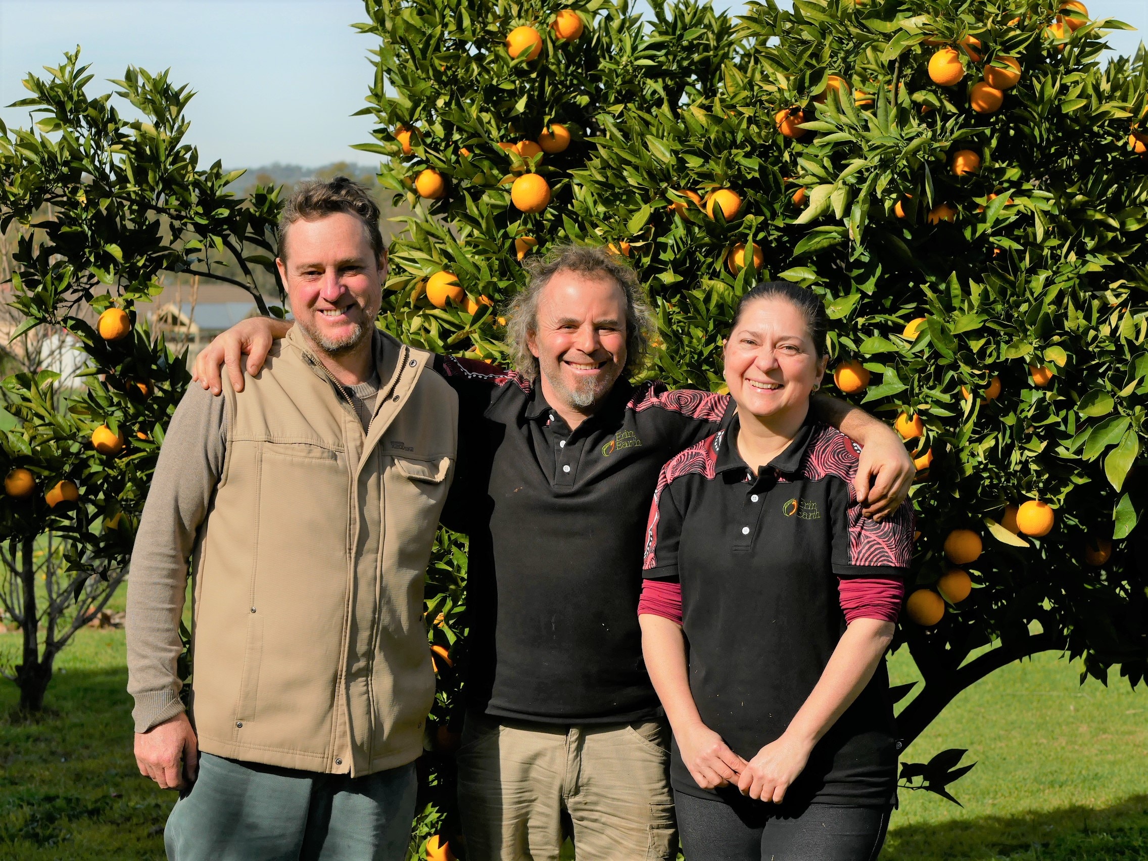 three people smiling in front of a orange tree.