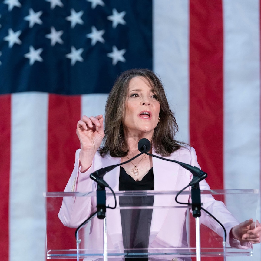 A slim older woman with long dark hair wearing a mauve suit speaks behind a clear lectern in front of a giant American flag.