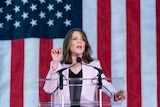 A slim older woman with long dark hair wearing a mauve suit speaks behind a clear lectern in front of a giant American flag.