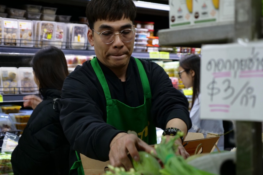 A man wearing a green apron handles green papayas at a grocery store