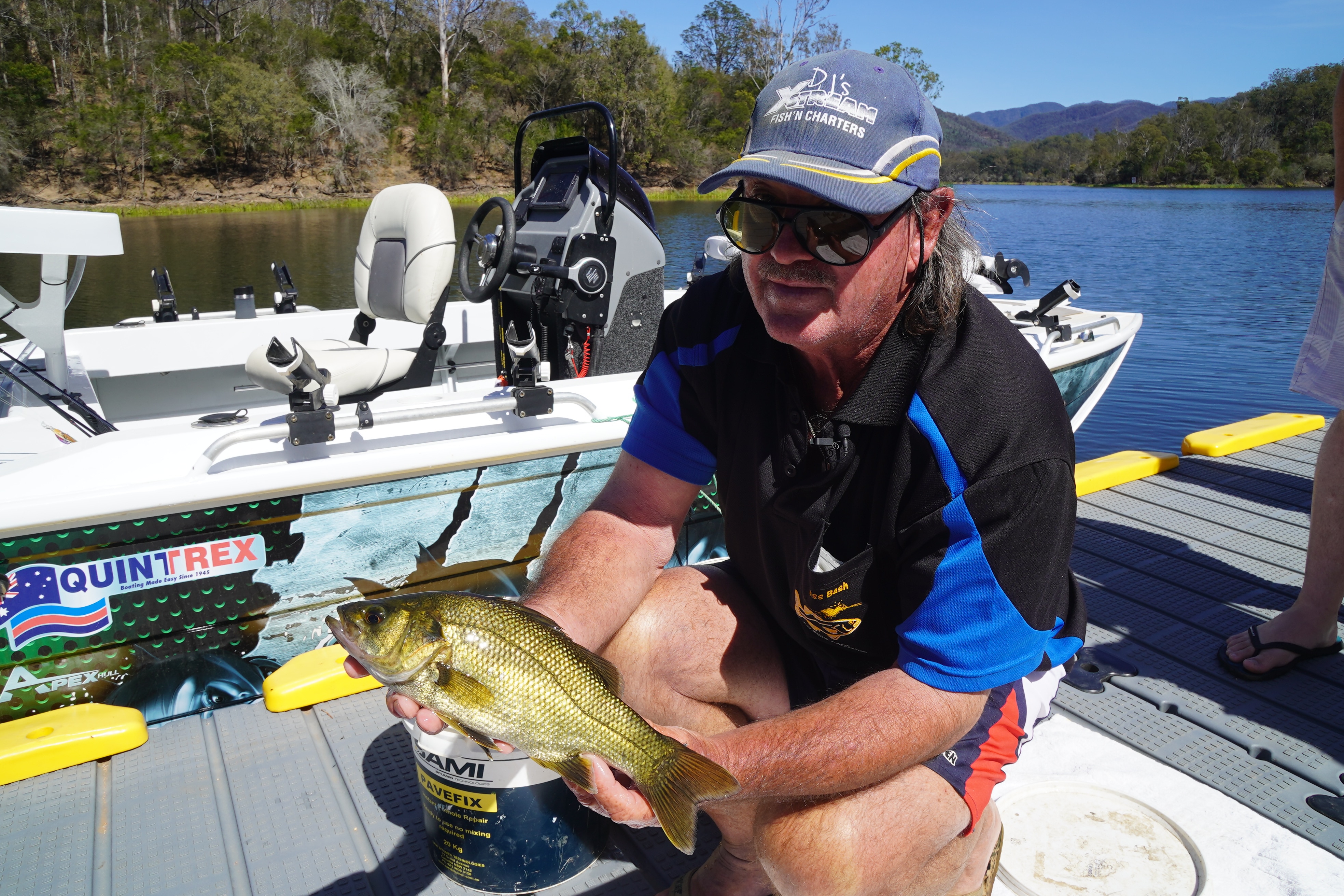 A man in a hat and glasses holds a bass fish in front of a dam.