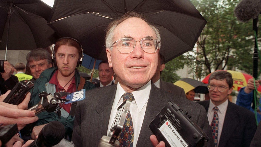 John Howard surrounded by microphones and media with umbrellas