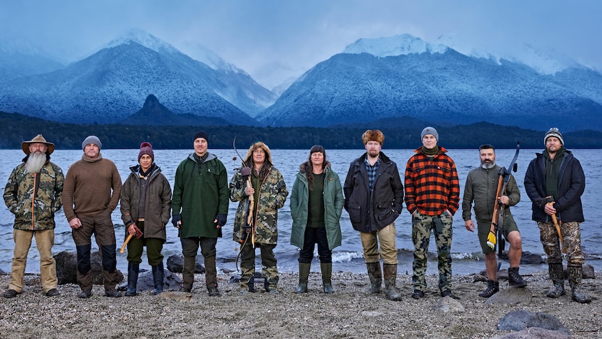 Ten people stand lined up by the shore of a lake with ice-capped mountains behind.