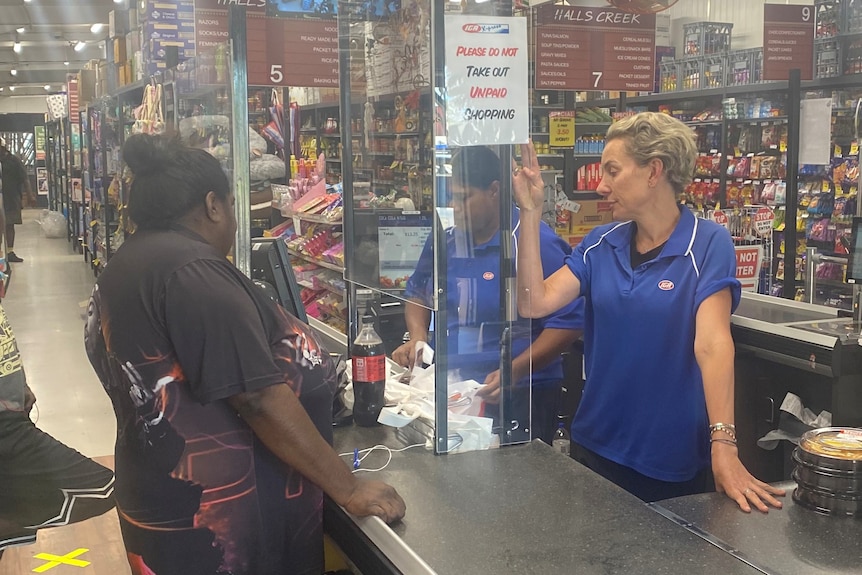 A blonde woman stands at a cash register with an Aboriginal woman next to her at an IGA store