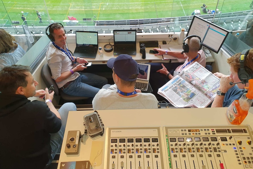 Five men sitting in a commentary box above a football ground.