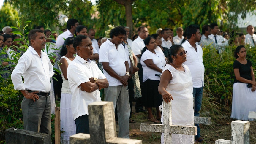 Men and women wear white and stand in a cemetery surrounded by crosses at graves.