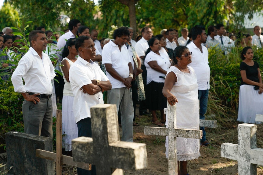 Men and women wear white and stand in a cemetery surrounded by crosses at graves.