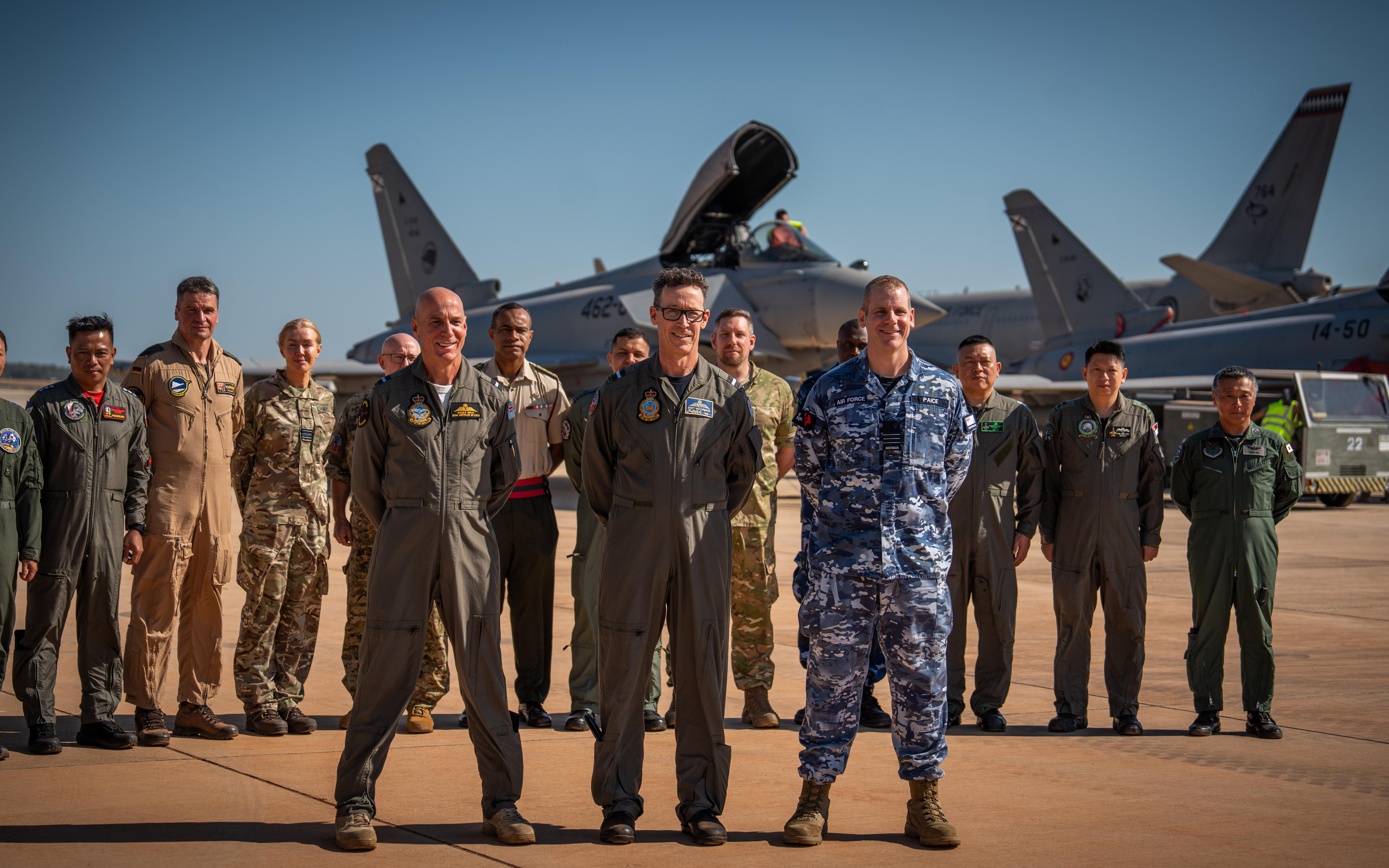 Three air force officers stand in front of other personnel with fighter jets in the background.