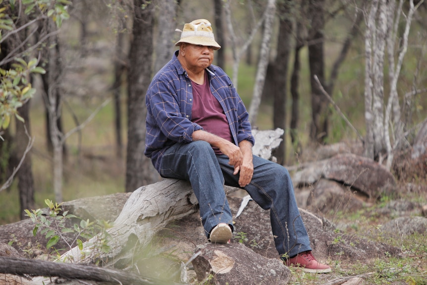 Indigenous man wearing a hat, shirt and jeans sitting on a fallen tree trunk.