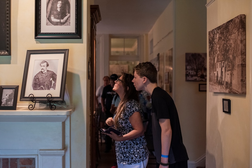 A man and a woman stand in the hallway of a home looking at portraits hung on a wall.