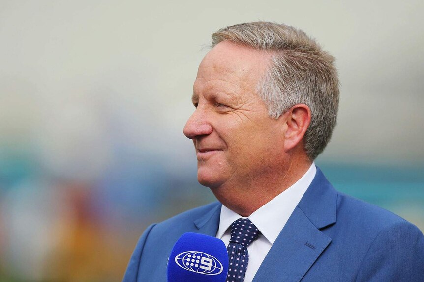 Australian cricketer Ian Healy, dressed in a blue suit, holds a microphone while commentating a match for Channel 9.