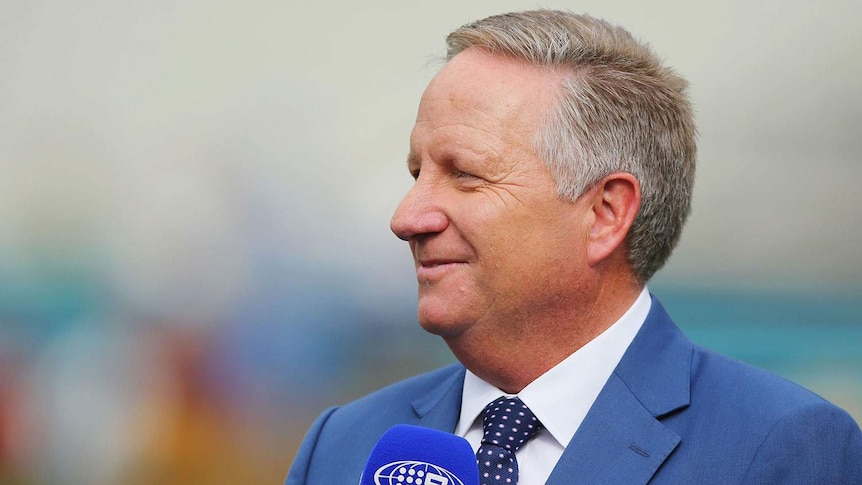 Australian cricketer Ian Healy, dressed in a blue suit, holds a microphone while commentating a match for Channel 9.