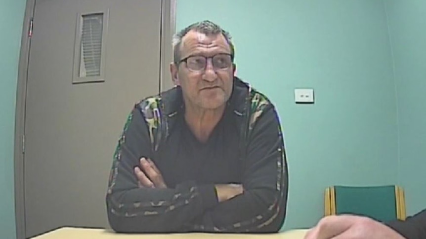 A man in a camouflage jacket with his arms crossed is spoken to in a police interview room 