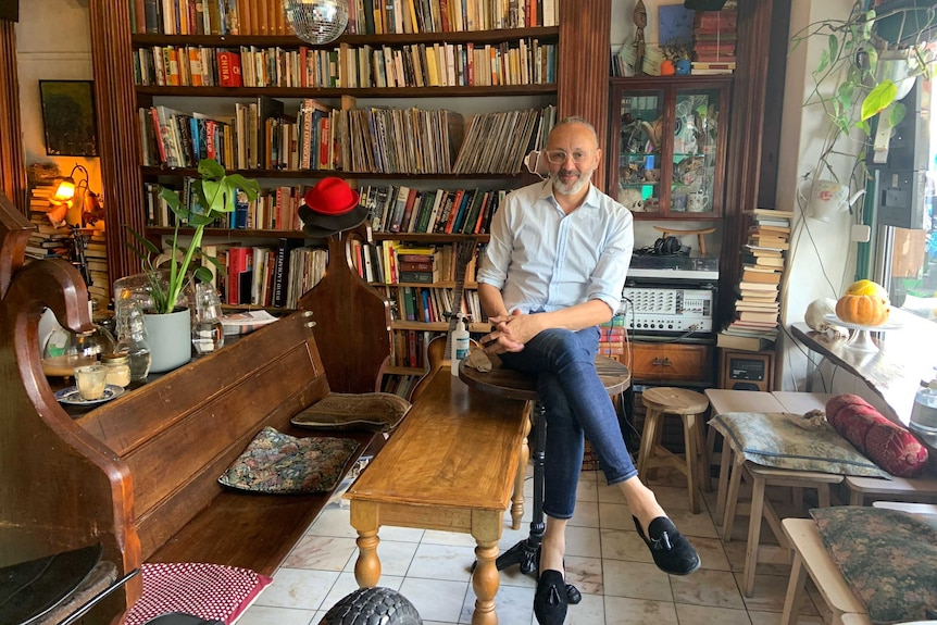 A man sits in a room with books behind him and surrounded by vintage chairs, tables and bric-a-brac