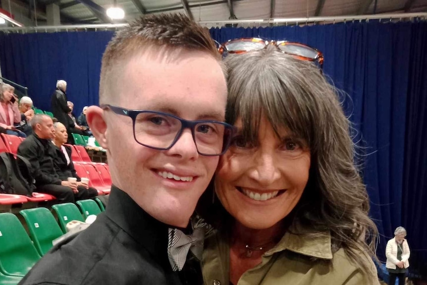 A young man in black shirt, glasses and bow tie is hugged by his mother in the stands of an event.