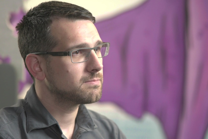 A man in glasses with short hair sits, a colourful painted wall behind him.