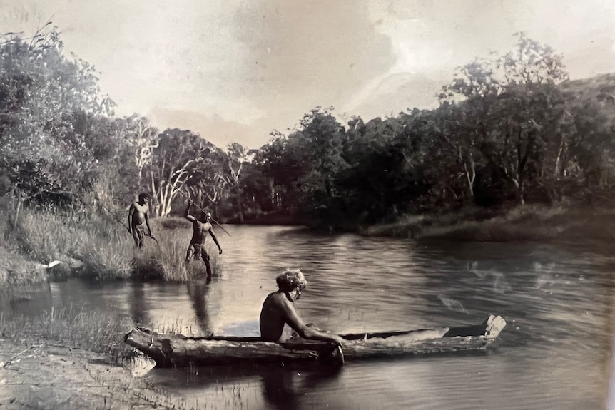An historical image of an Aboriginal man in a traditional canoe with two other men on the river bank, all without clothes.