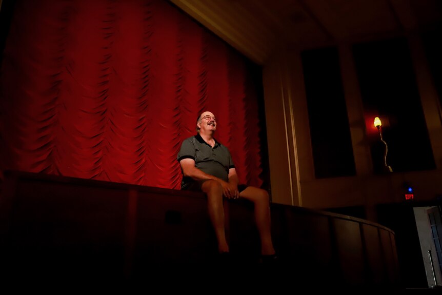 Man sits on edge of theatre stage with large red curtain behind him. 