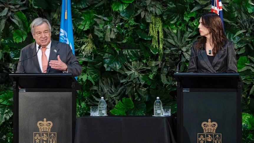 Antonio Guterres stands on a podium next to Jacinda Ardern.