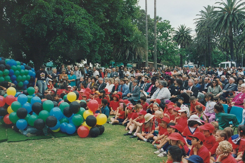 View of audience, including primary school children, with blue, black, red, yellow and green balloons.
