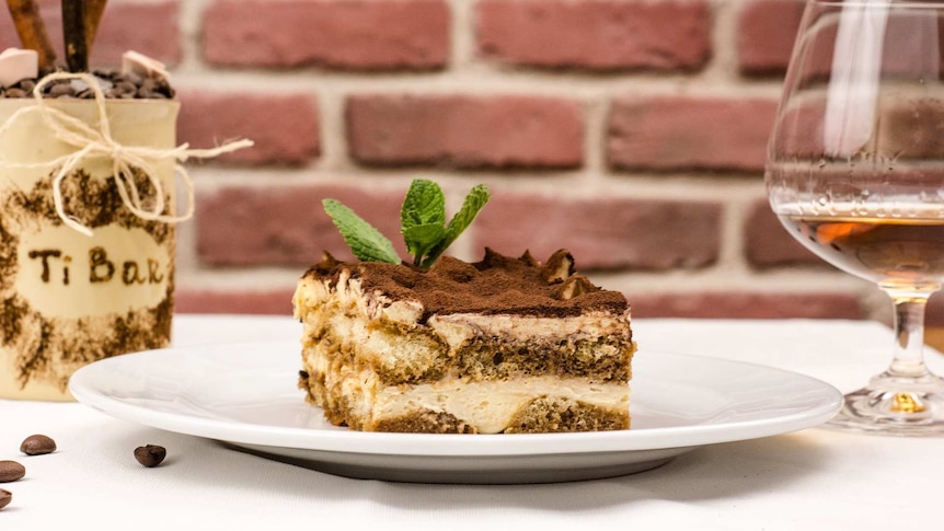 A slice of tiramisu on a white plate, with a glass of liqueur to its right.