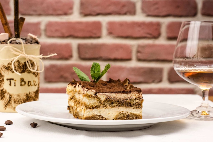 A slice of tiramisu on a white plate, with a glass of liqueur to its right.