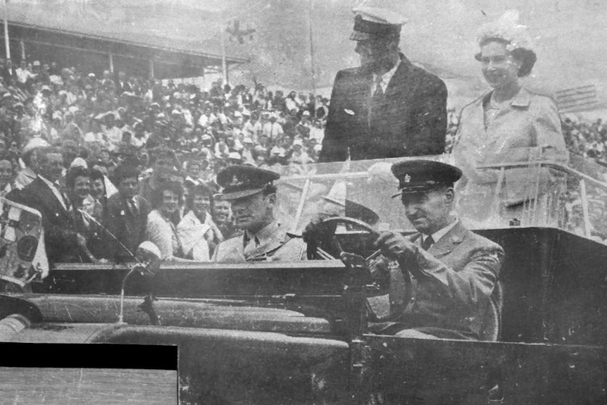 Black and white photo of the Queen and Prince Philip standing in the back of a car with a crowd of people watching on