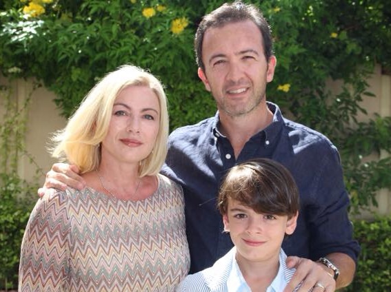 A woman, man and young boy standing in the sun in front of a fence with a bush with yellow flowers on it