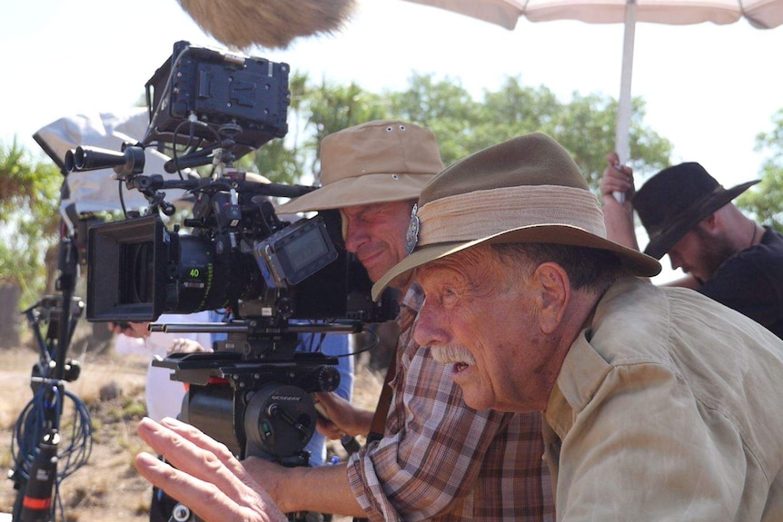 An older man wearing an army uniform stands next to a camera