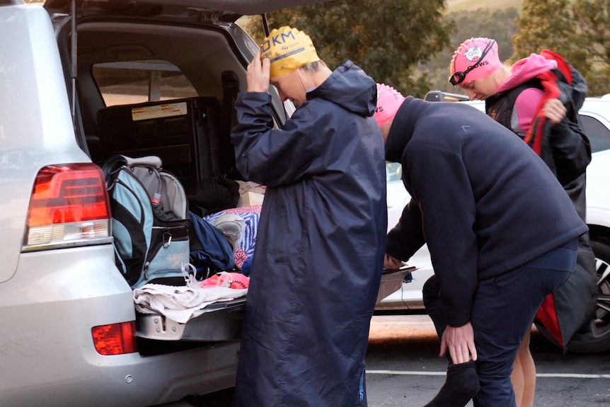 Three people in overcoats and swim caps prepare to undress at their car boots in the dark winter dawn
