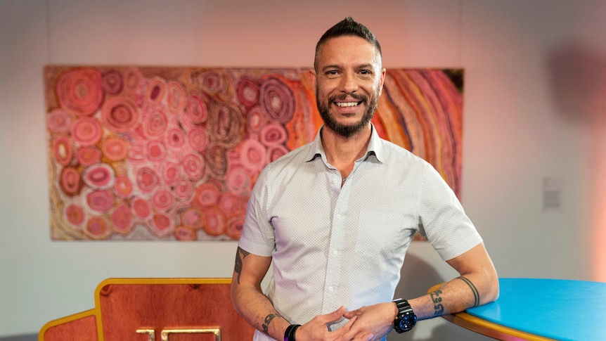An Aboriginal man with short hair, beard and white shirt standing in front a colourful Aboriginal dot painting