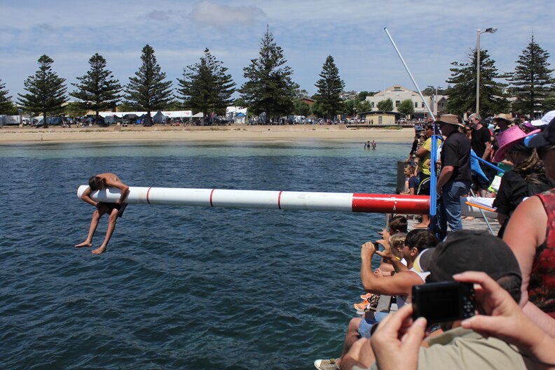 A crowd standing on a jetty watches a boy hanging from a pole over the water as part of the slippery pole competition.