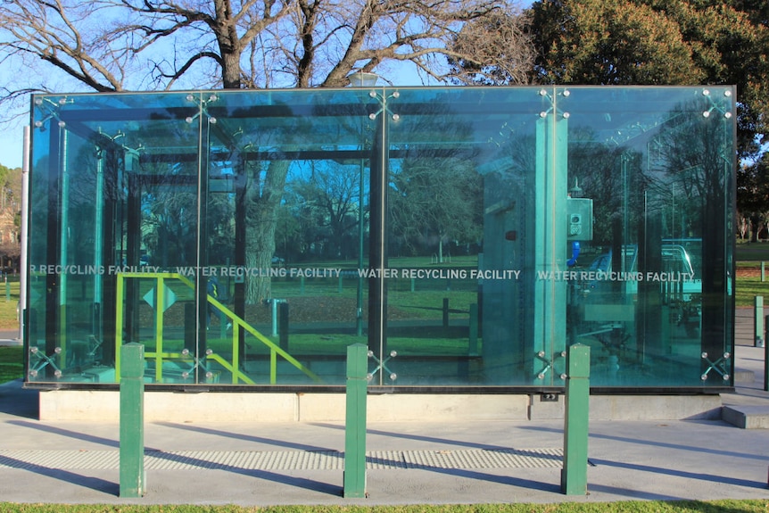 A blue glass cube with the words water recycling facility in a park with an internal staircase leading underground.