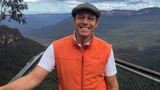 Ian Randall stands in front of a valley wearing an orange Gillet and grey flat cap