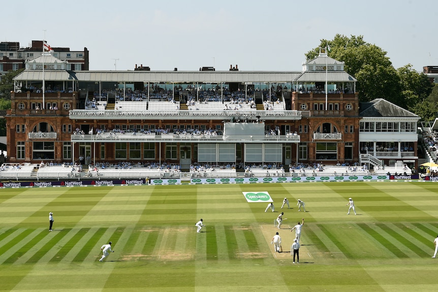 An old pavilion style building and a large green field with cricketers planing cricket. 