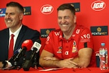 Trevor Gleeson smiles at a press conference, sitting at a table with microphones wearing a red shirt.