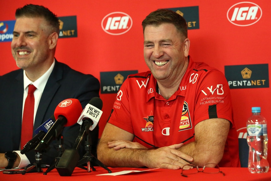 Trevor Gleeson smiles at a press conference, sitting at a table with microphones wearing a red shirt.