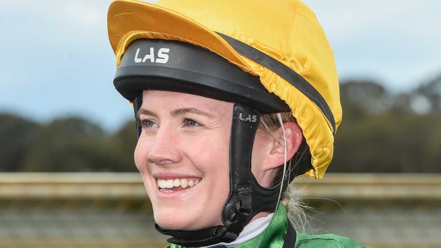 Mikaela Claridge smiles, dressed in her jockey's outfit, helmet and yellow hat.