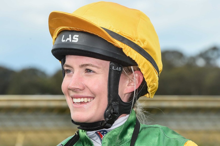 Mikaela Claridge smiles, dressed in her jockey's outfit, helmet and yellow hat.
