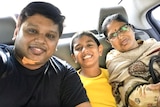 A man wearing a black shirt smiles in the back seat of a car next to a boy wearing a yellow shit and a woman wearing a shawl.