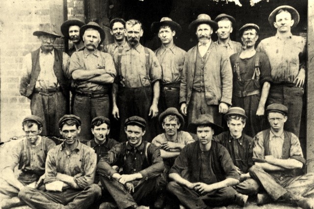Black and white photo of miners standing and sitting. Most wear hats and dirty clothes.